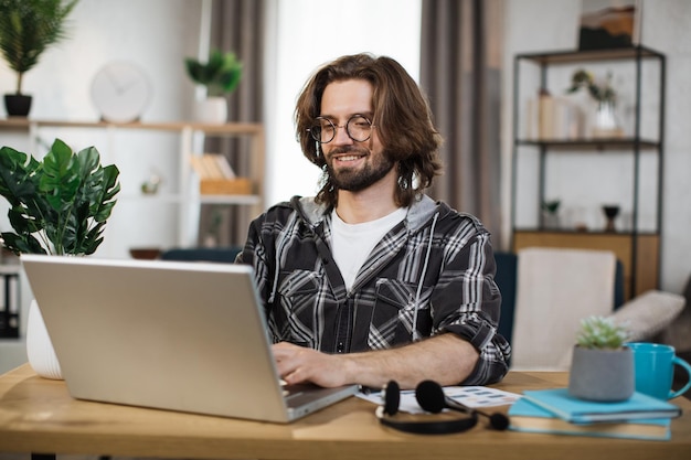 Uomo caucasico barbuto con gli occhiali in abiti casual che lavora su un computer portatile wireless mentre è seduto alla scrivania