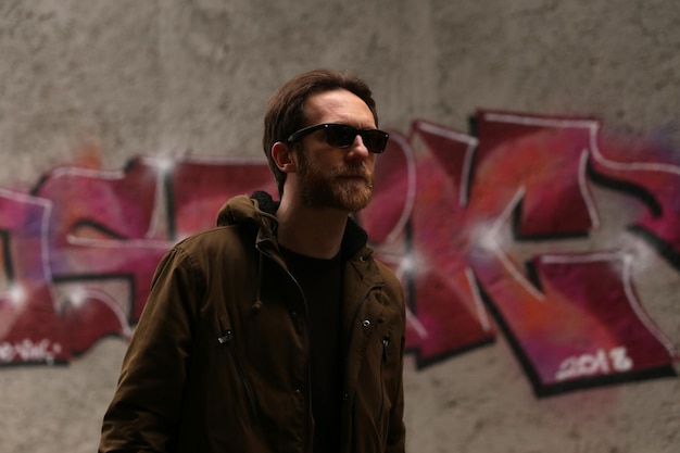 Uomo caucasico barbuto con occhiali da sole e giacca marrone in posa davanti a un graffito