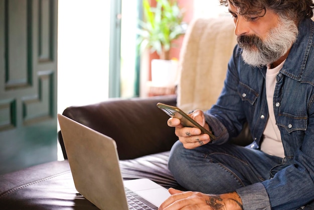 Uomo caucasico barbuto di mezza età che utilizza laptop e smart phonelavoro remoto social media a casa