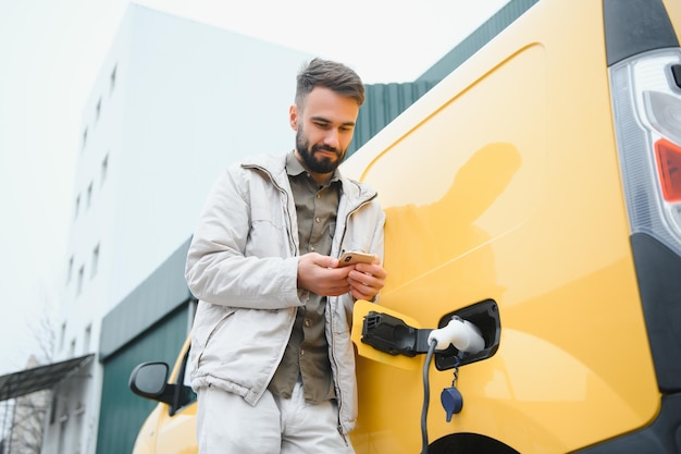 Uomo caucasico barbuto in piedi vicino a un'auto elettrica che sta caricando e regolando il tempo su uno smartphone