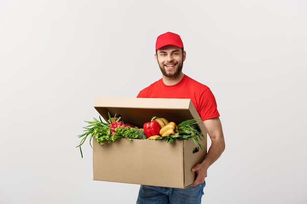 Uomo caucasico bello del corriere di consegna della drogheria in uniforme rossa con il contenitore di drogheria con frutta e la verdura fresche