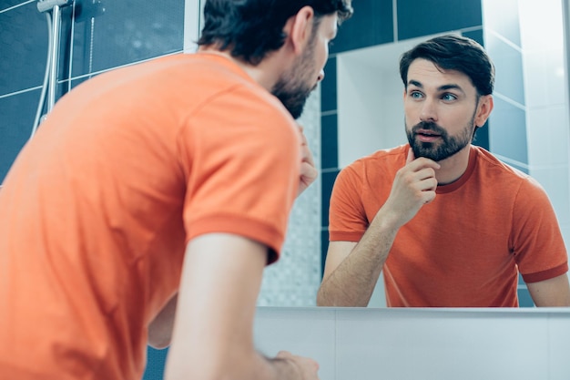 Uomo caucasico calmo che si tocca la barba e guarda il riflesso del suo viso nello specchio del bagno