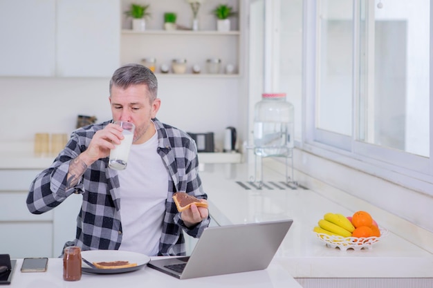 Uomo caucasico che beve latte mangiando colazione mentre lavora online sul computer portatile