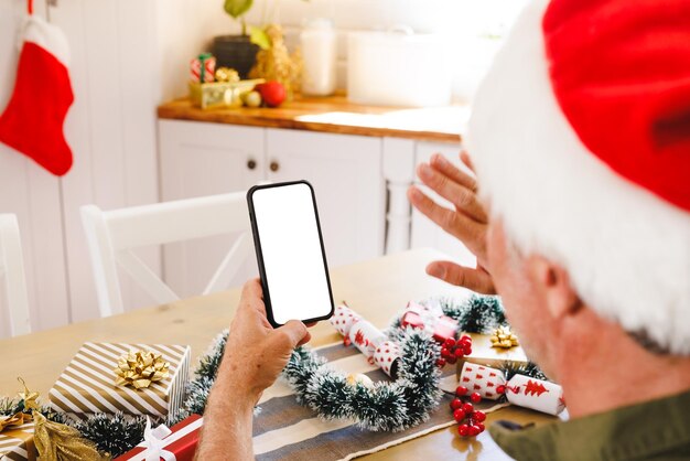 Uomo caucasico che indossa un cappello da Babbo Natale, seduto al tavolo in cucina, utilizzando uno smartphone con copyspace