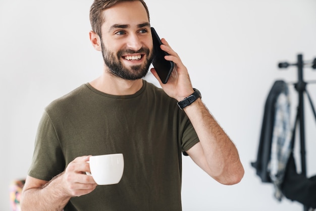 uomo caucasico che ride che indossa una maglietta casual che parla al cellulare e beve caffè mentre lavora in un ufficio luminoso