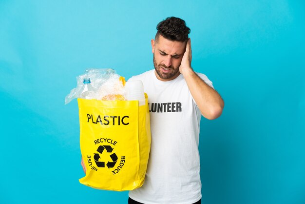 Uomo caucasico che tiene un sacchetto pieno di bottiglie di plastica da riciclare isolato su sfondo blu con mal di testa