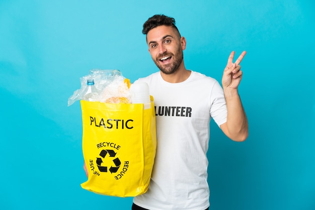 Uomo caucasico che tiene un sacchetto pieno di bottiglie di plastica da riciclare isolato su sfondo blu sorridendo e mostrando il segno della vittoria