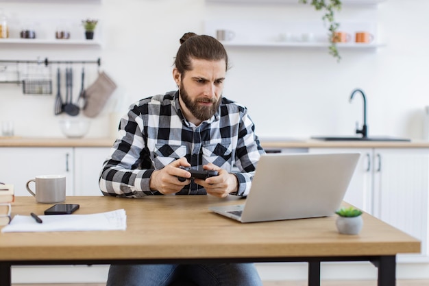 Uomo caucasico che usa un joystick e un portatile seduto a un tavolo di legno in cucina