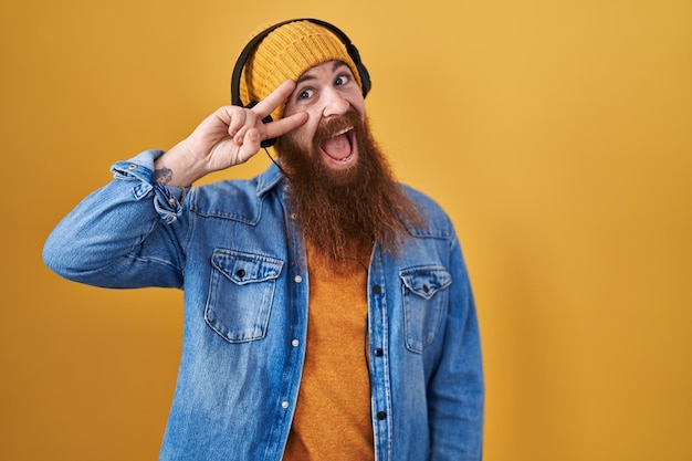 Uomo caucasico con barba lunga che ascolta musica utilizzando le cuffie facendo simbolo di pace con le dita sul viso sorridente allegro mostrando vittoria