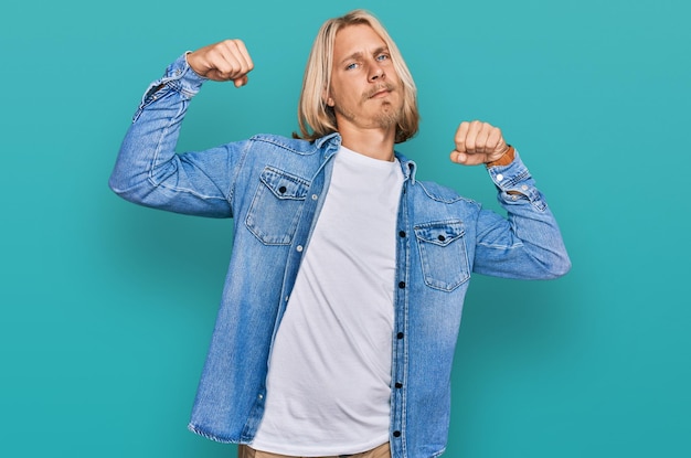 Uomo caucasico con capelli lunghi biondi che indossa una giacca di jeans casual che mostra i muscoli delle braccia sorridendo fiero concetto di fitness