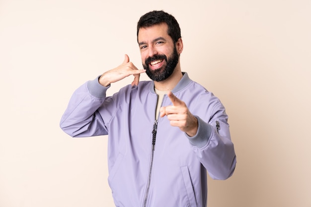 Uomo caucasico con la barba che indossa una giacca sopra la parete isolata che fa il gesto del telefono e che indica la parte anteriore