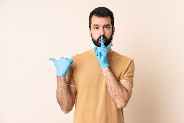 Uomo caucasico con la barba che protegge con una maschera e guanti sopra la parete che indica il lato e che fa gesto di silenzio