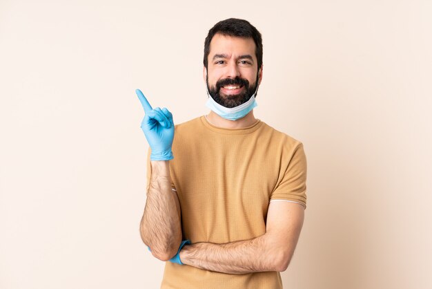 Uomo caucasico con la barba che protegge con una maschera e guanti sopra la parete felice e che indica in su