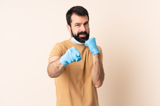 Uomo caucasico con la barba che protegge dal coronavirus con una maschera e guanti sopra la parete con il gesto di combattimento