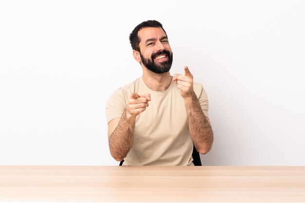 Uomo caucasico con la barba in un tavolo che punta davanti con felice espressione.