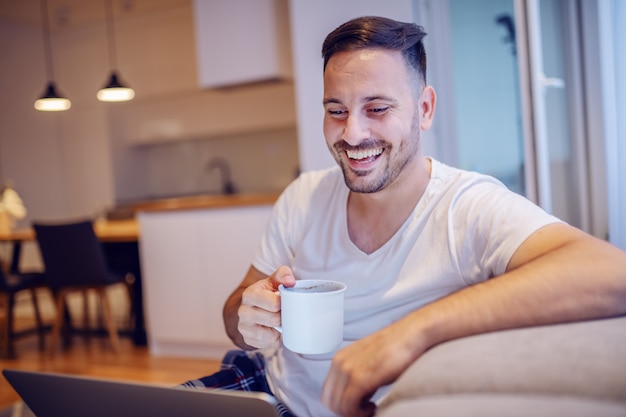 Uomo caucasico con la barba lunga sorridente bello in pigiama che si siede nel salone con il computer portatile in grembo e che beve il suo caffè fresco di mattina.