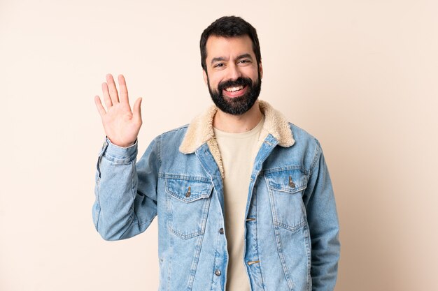 Uomo caucasico con la barba sopra la parete isolata che saluta con la mano con l'espressione felice