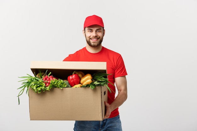 Uomo caucasico del corriere di consegna della drogheria in uniforme rossa con la scatola di drogheria con frutta fresca