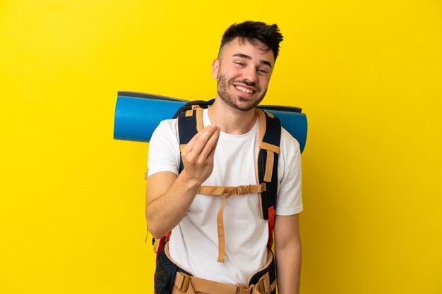 Uomo caucasico del giovane alpinista con un grande zaino isolato su fondo giallo che fa il gesto dei soldi