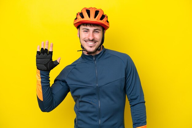 Uomo caucasico del giovane ciclista isolato su fondo giallo che saluta con la mano con l'espressione felice