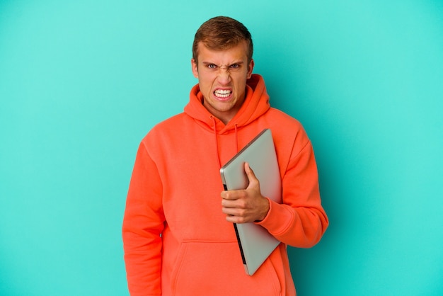 Uomo caucasico del giovane studente che tiene un computer portatile isolato sul blu che grida molto arrabbiato e aggressivo.