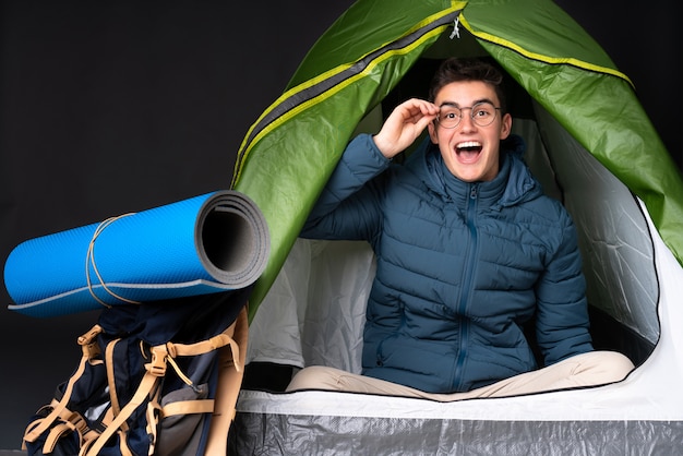 Uomo caucasico dell'adolescente dentro una tenda verde di campeggio isolata sul nero con i vetri e sorpreso