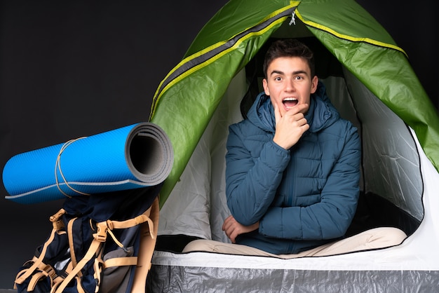 Uomo caucasico dell'adolescente dentro una tenda verde di campeggio isolata sul nero sorpreso e colpito mentre sembrando giusto