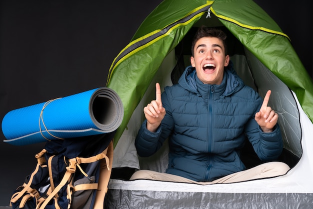 Uomo caucasico dell'adolescente dentro una tenda verde di campeggio sulla parete nera sorpresa e che indica su