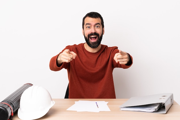 Uomo caucasico dell'architetto con la barba in un tavolo sorpreso e indicando la parte anteriore.