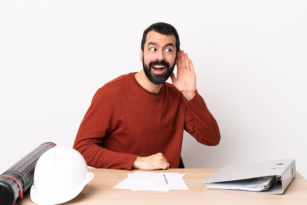 Uomo caucasico dell'architetto con la barba in una tabella che ascolta qualcosa mettendo la mano sull'orecchio.