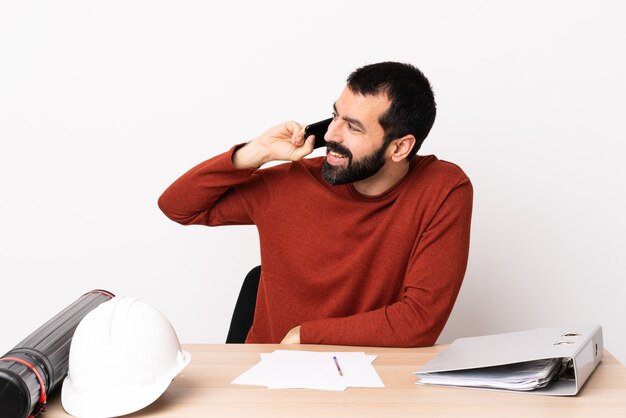 Uomo caucasico dell'architetto con la barba in una tabella che mantiene una conversazione con il telefono cellulare con qualcuno.