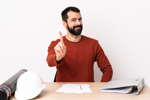 Uomo caucasico dell'architetto con la barba in una tabella che mostra e che alza un dito.