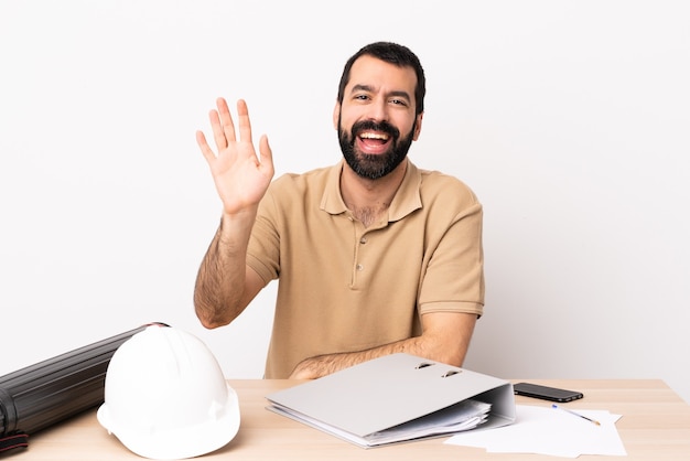 Uomo caucasico dell'architetto con la barba in una tabella che saluta con la mano con l'espressione felice.