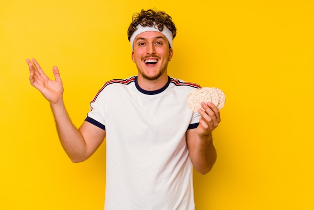 Uomo caucasico di giovane sport che mangia una torta di riso isolata su priorità bassa gialla che riceve una sorpresa piacevole, eccitata e alzando le mani.