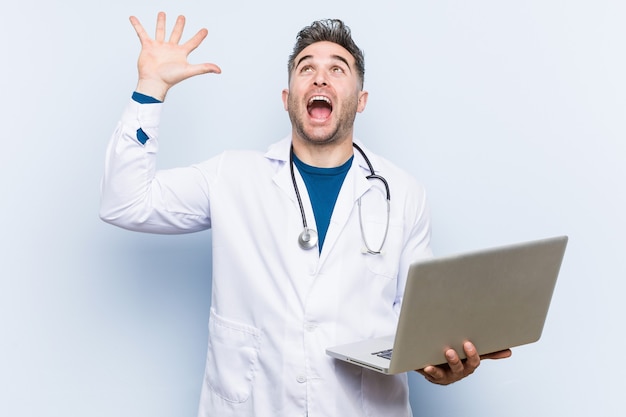 Uomo caucasico di medico che tiene un computer portatile che celebra una vittoria o un successo