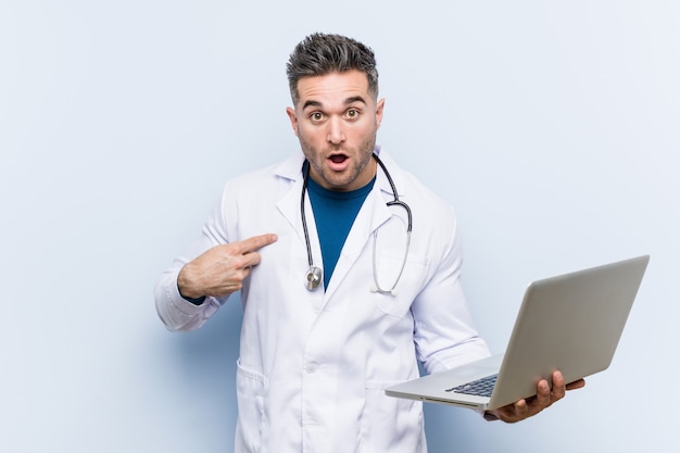 Uomo caucasico di medico che tiene un computer portatile sorpreso indicando se stesso, sorridendo ampiamente.