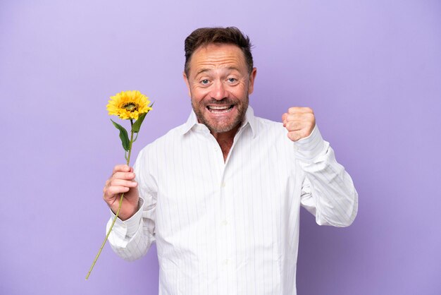 Uomo caucasico di mezza età che tiene il fiore del sole isolato su sfondo viola che celebra una vittoria nella posizione del vincitore