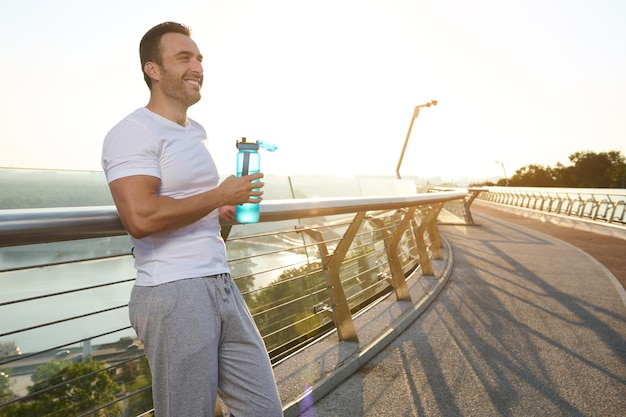 Uomo caucasico di mezza età in forma muscoloso sportivo atleta che beve acqua mentre ci si rilassa dopo la corsa mattutina sul ponte della città Concetto di stile di vita attivo sano di reidratazione