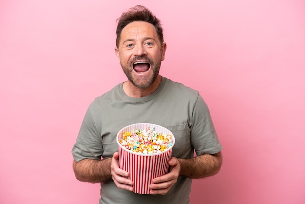 Uomo caucasico di mezza età isolato su sfondo rosa con in mano un grande secchio di popcorn