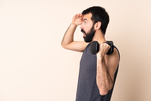 Uomo caucasico di sport con la barba che fa sollevamento pesi sopra fondo isolato con espressione di sorpresa mentre guarda lato