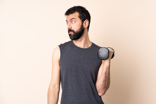 Uomo caucasico di sport con la barba che fa sollevamento pesi sopra isolato facendo dubbi gesto guardando lato