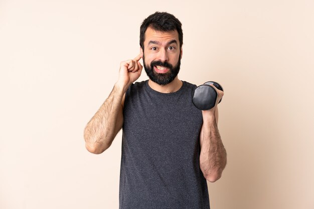 Uomo caucasico di sport con la barba che fa sollevamento pesi sopra la parete frustrata e che copre le orecchie