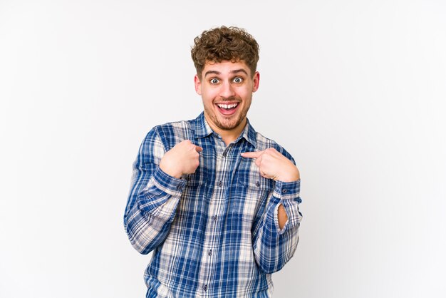 Uomo caucasico isolato dei giovani capelli ricci biondi isolato indicare sorpreso con la barretta, sorridente ampiamente.