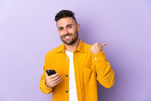 Uomo caucasico isolato sulla parete viola utilizzando il telefono cellulare e indicando il laterale