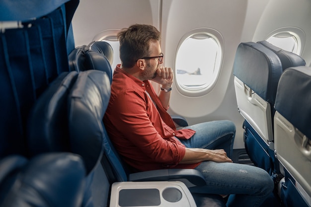 Uomo caucasico pensieroso in abbigliamento casual che riposa sull'aereo, seduto e guardando fuori dalla finestra. Relax, viaggio, vacanza, concetto di trasporto