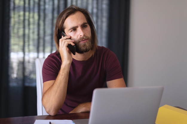Uomo caucasico seduto al tavolo che lavora da casa e parla sullo smartphone