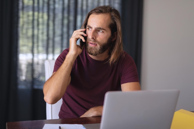 Uomo caucasico seduto al tavolo che parla sullo smartphone e lavora da casa