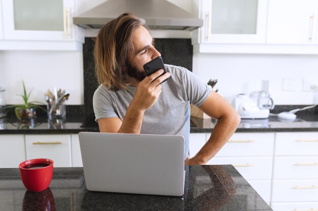 Uomo caucasico seduto in cucina utilizzando laptop e smartphone