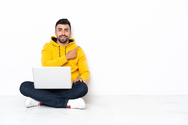 Uomo caucasico seduto sul pavimento con il suo laptop rivolto di lato per presentare un prodotto