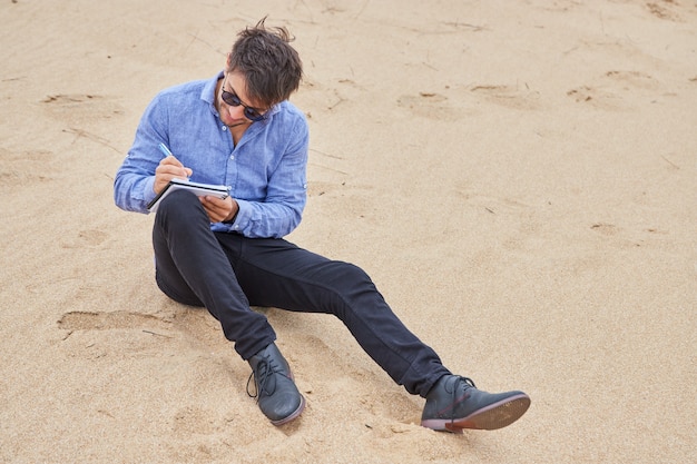 Uomo caucasico seduto sulla sabbia sulla spiaggia che scrive poesie concentrate o una lettera. Indossa occhiali da sole, maglietta blu, pantaloni neri e scarpe nere.